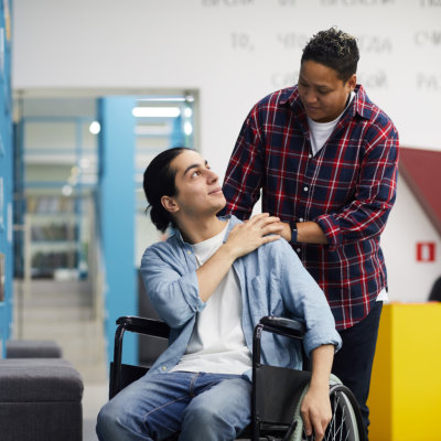 Portrait of two college students, one of them in wheelchair, looking at each other while standing by shelves in library, copy space